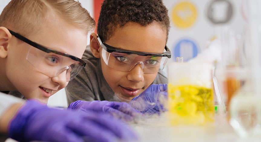 boys in safety goggles leaning closely to the table and watching chemical reaction going on in the beaker