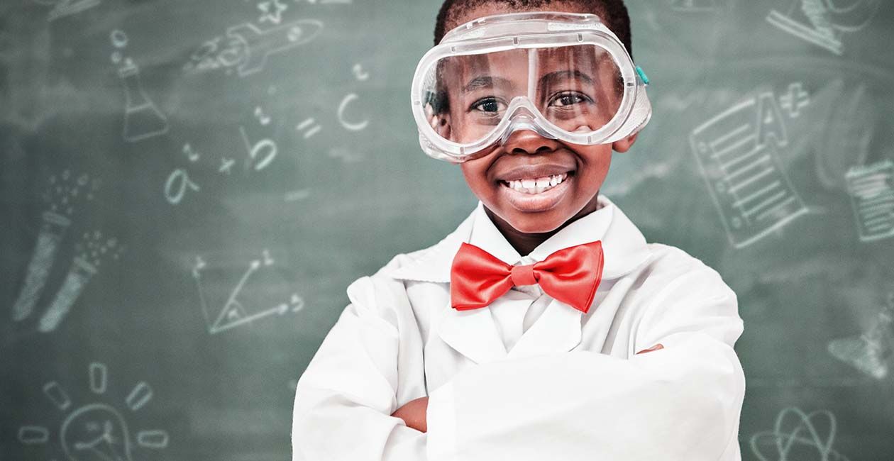 Boy wearing lab coat, red bow tie and safety goggles in a classroom.  Blackboard in the background with different science ico