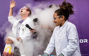 Mad scientist holding a microphone behind a cloud of smoke in between a girl and mad scientist both in lab coats
