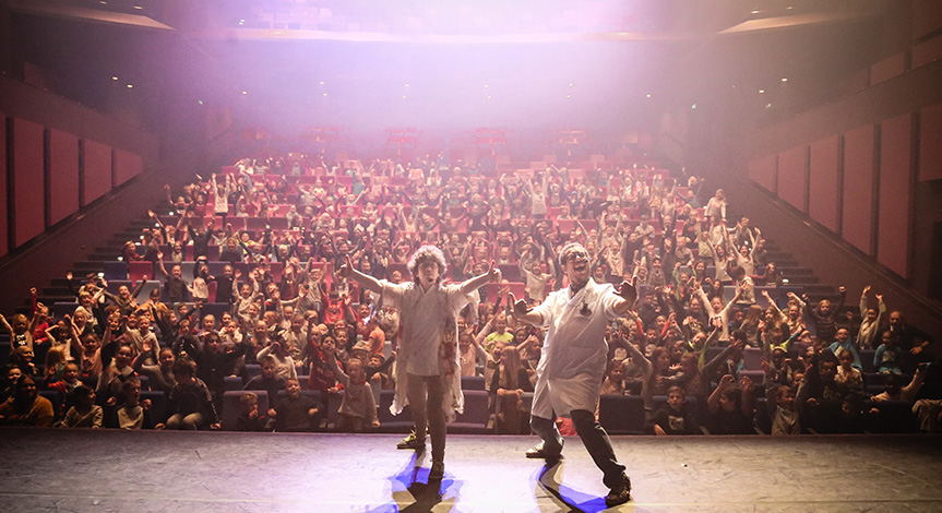 2 Mad scientists in labcoats on stage with their back towards a large excited crowd 