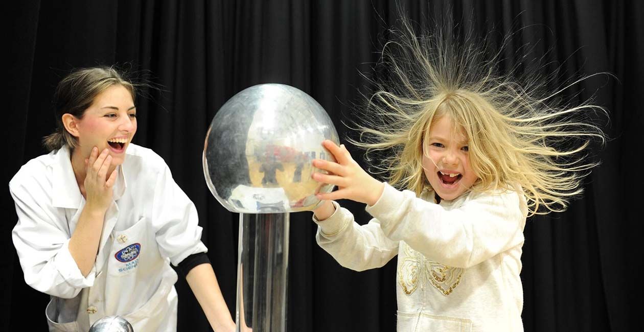 Girl touching a plasma ball causing her hair to to stand up while her mad science instructor looks on.