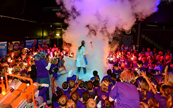 Mad scientist performing on stage surrounded by kids standing in a cloud of smoke